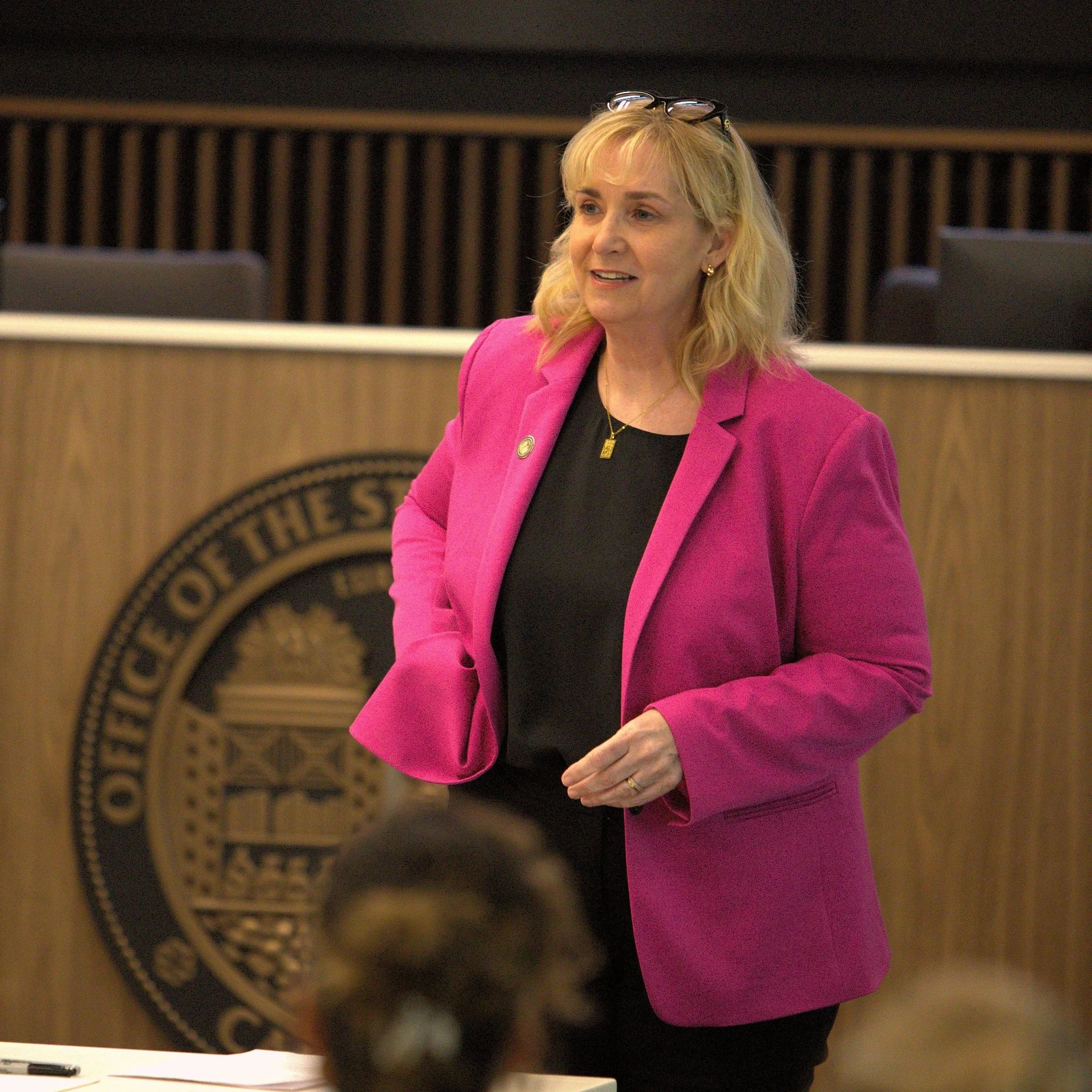 A woman with blonde hair, glasses on her head, wearing a pink blazer and gold jewelry, speaking in a formal setting with official seals or emblems in the background.