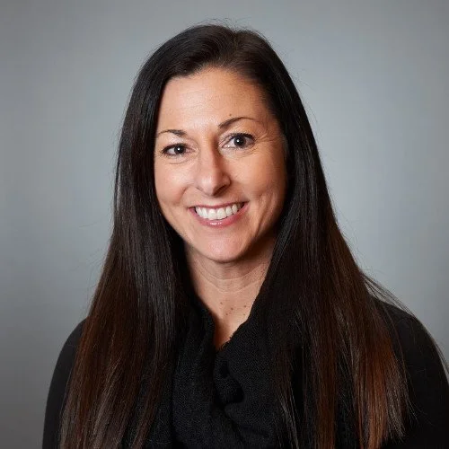 Photograph of a woman with long dark hair smiling against a neutral gray background.