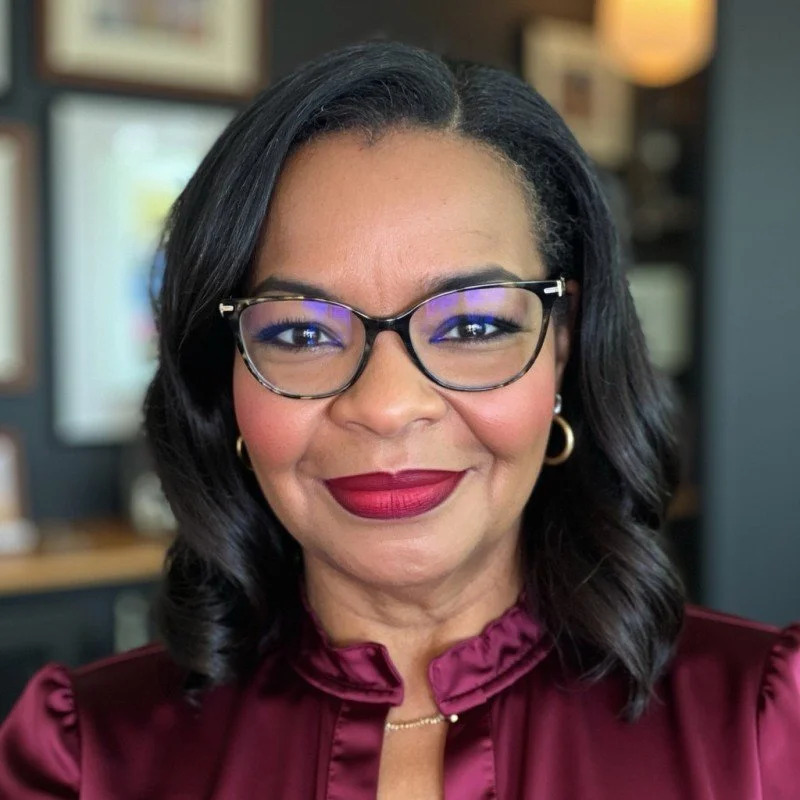 A woman with black hair, glasses, maroon lipstick, gold earrings, and a maroon blouse smiling indoors.