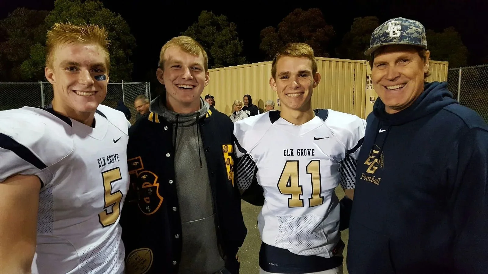 Four young men and an adult man smiling for a photo on a football field at night. The young men are wearing white football jerseys with 'ELK GROVE' and numbers. The adult man is wearing a hat and a navy hoodie with the 'ELK GROVE Football' logo.