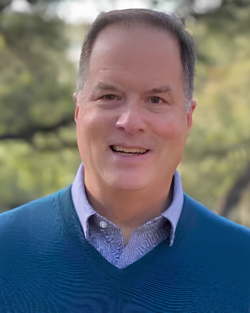 A smiling man with short dark hair, wearing a blue sweater over a collared shirt, standing outdoors with blurred trees in the background.