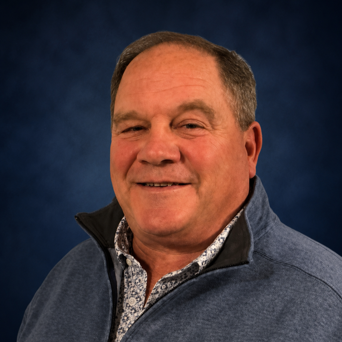 A middle-aged man smiling, wearing a blue zip-up sweater over a floral collared shirt, with a dark blue background.
