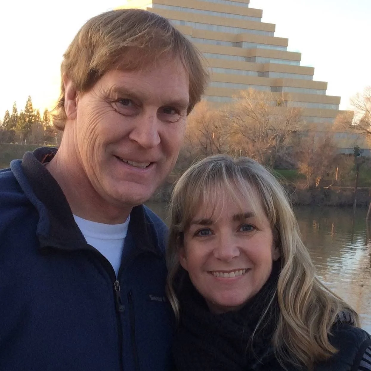 A smiling man and woman taking a selfie outdoors near a body of water, with a modern building and trees in the background during sunset.