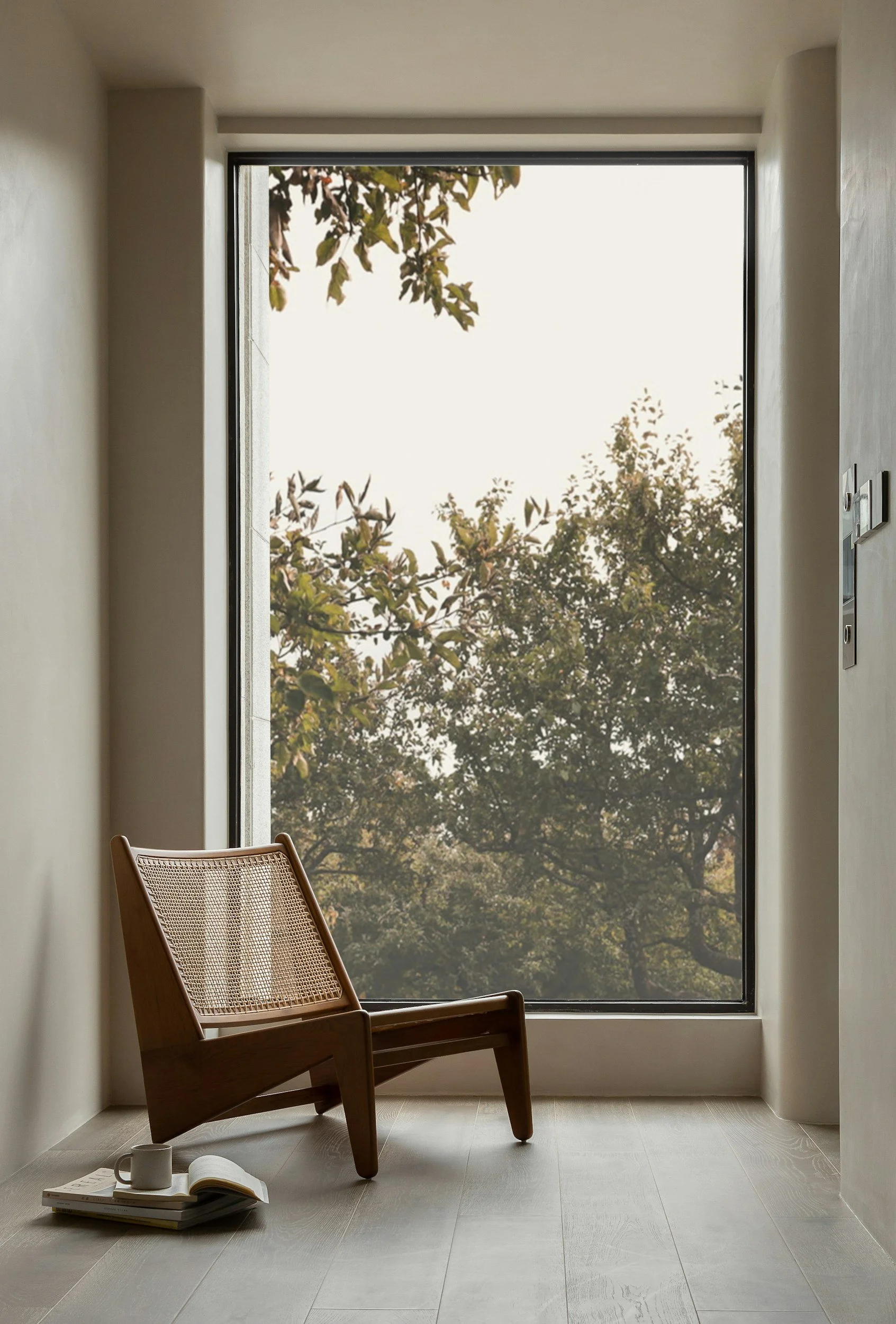 A modern interior with a large floor-to-ceiling window showing trees outside. A wooden chair with a woven seat is positioned near the window, and a small stack of books, a cup, and an open notebook are on the floor.
