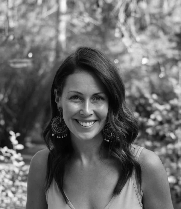 A black and white photo of a woman with shoulder-length wavy hair smiling outdoors, wearing large circular earrings and a sleeveless top, with a blurred natural background.
