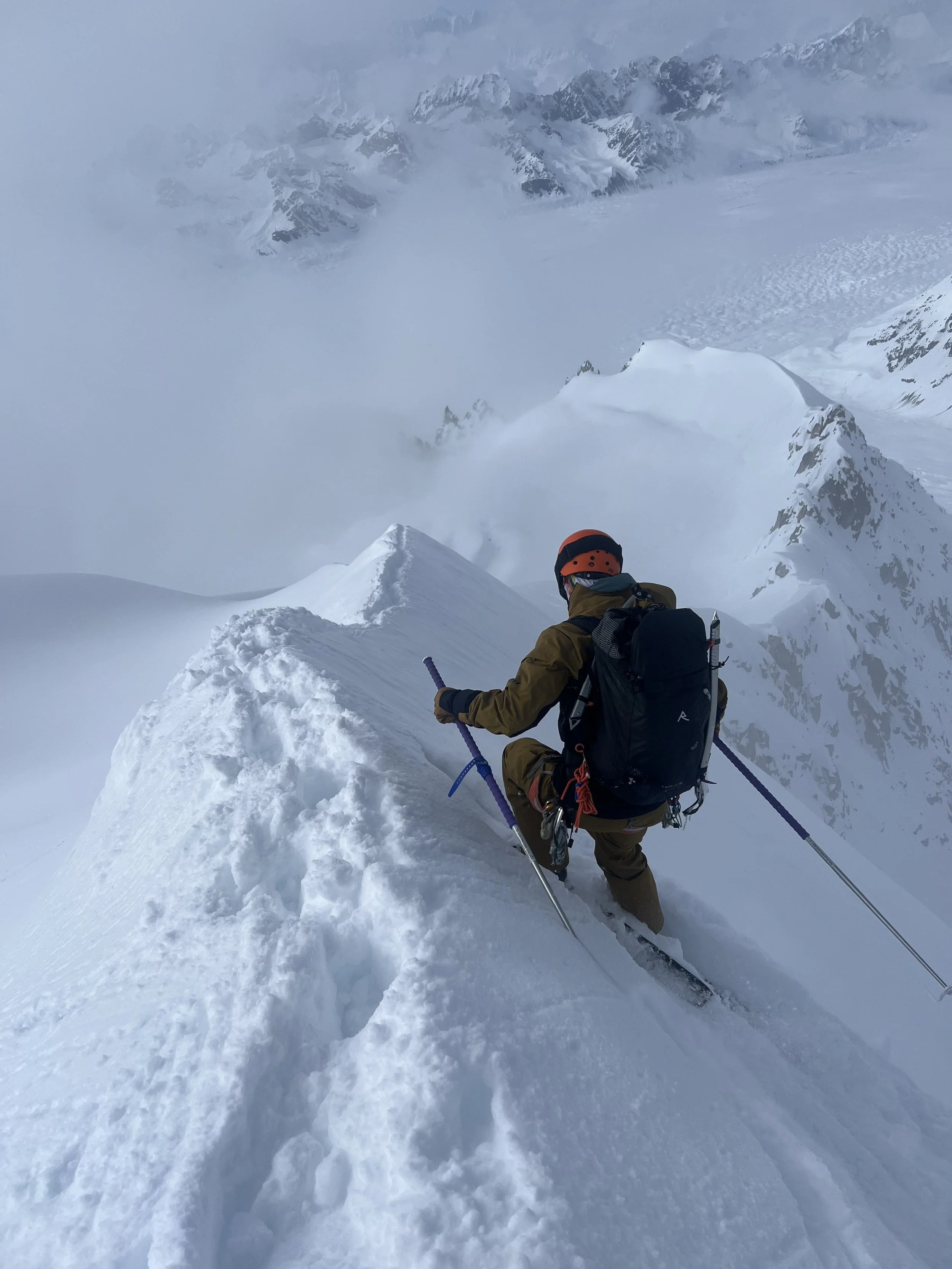 A mountaineer wearing a helmet and winter gear ascending a steep snowy mountain ridge, surrounded by snow-covered peaks and cloudy sky.