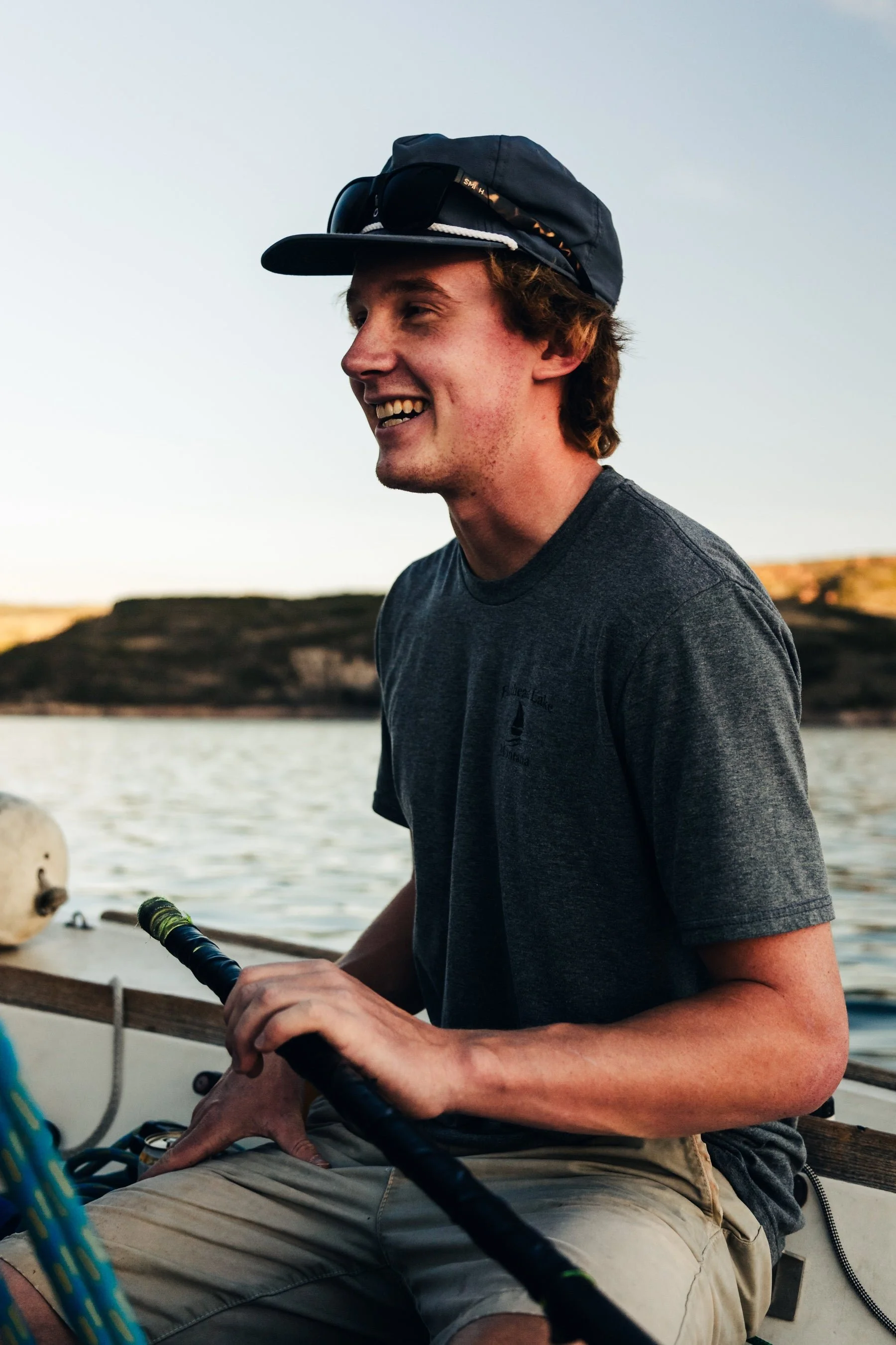 Young man with light skin, curly hair, wearing a dark cap with sunglasses and a dark t-shirt, smiling while holding a fishing rod on a boat, with a body of water and land in the background.