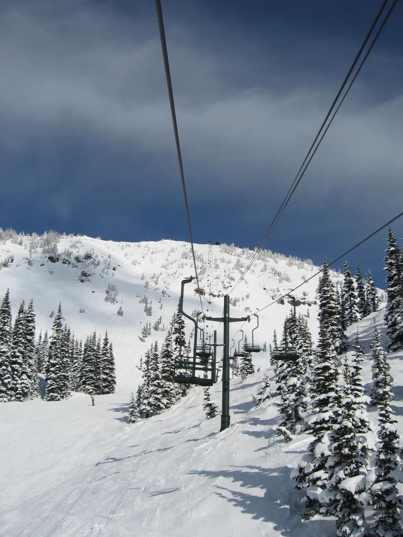 Snow-covered mountain with ski lift in front of trees, under a cloudy sky.