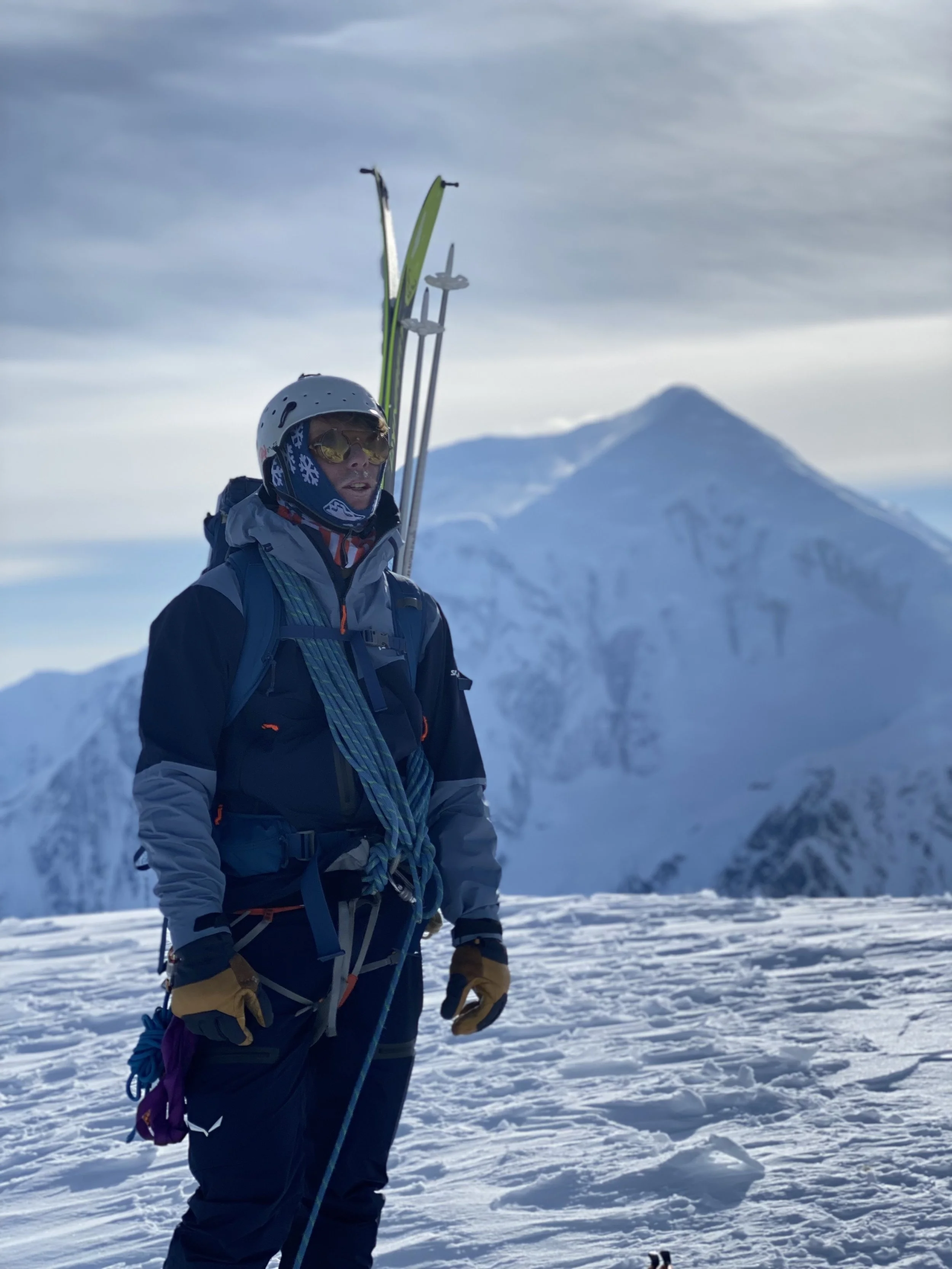A person dressed in winter gear with skis and ski poles, standing on snow with a snow-covered mountain in the background.