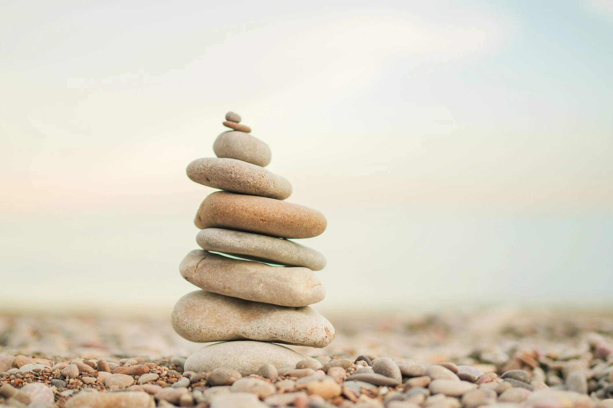 Stack of smooth, rounded stones balanced on top of each other on a beach, with a blurry sandy background and a pale sky.