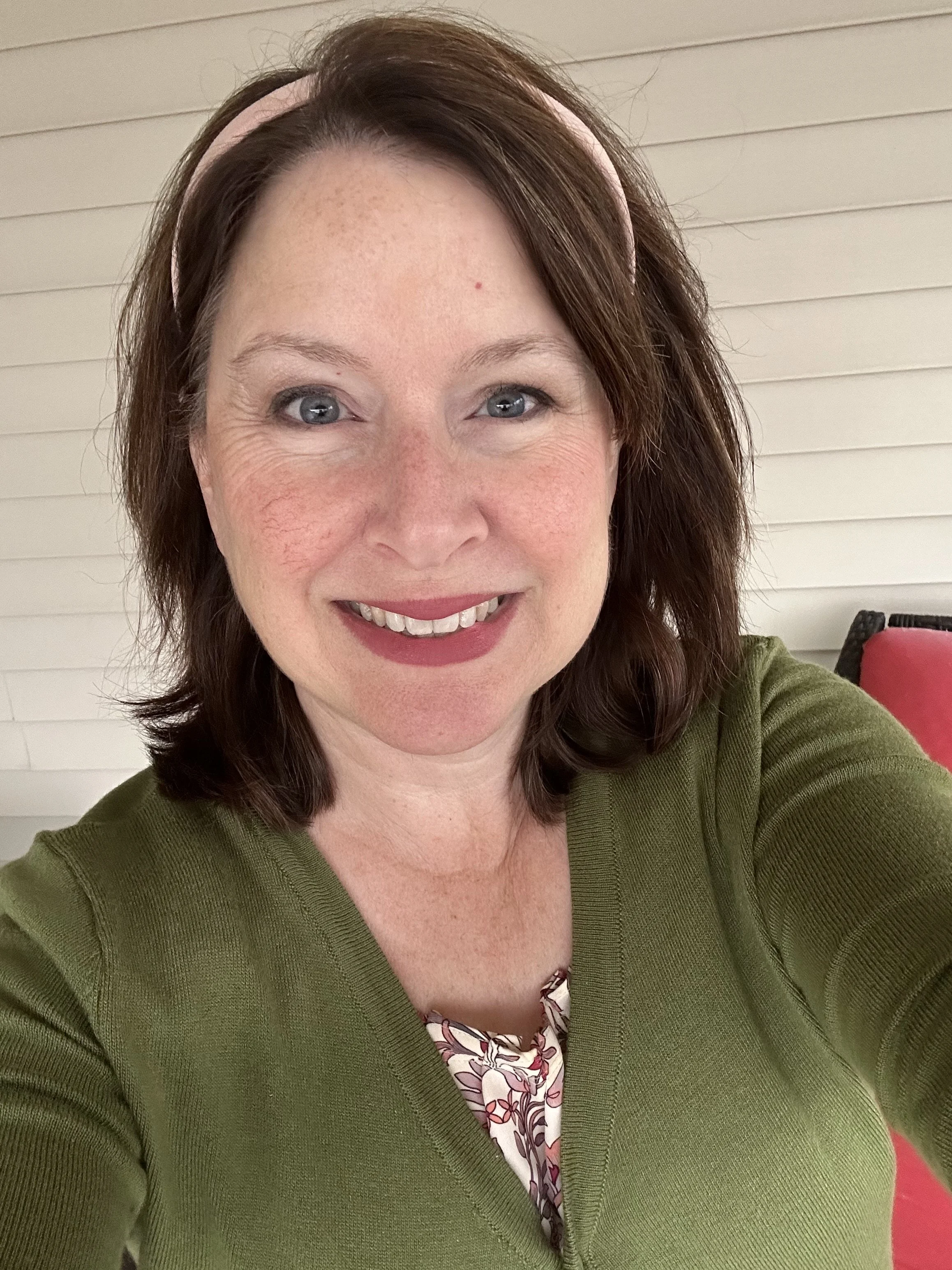 Close-up of a woman smiling, wearing a green sweater, with a light pink headband, and sitting in an indoor setting with beige shiplap walls and a red chair in the background.