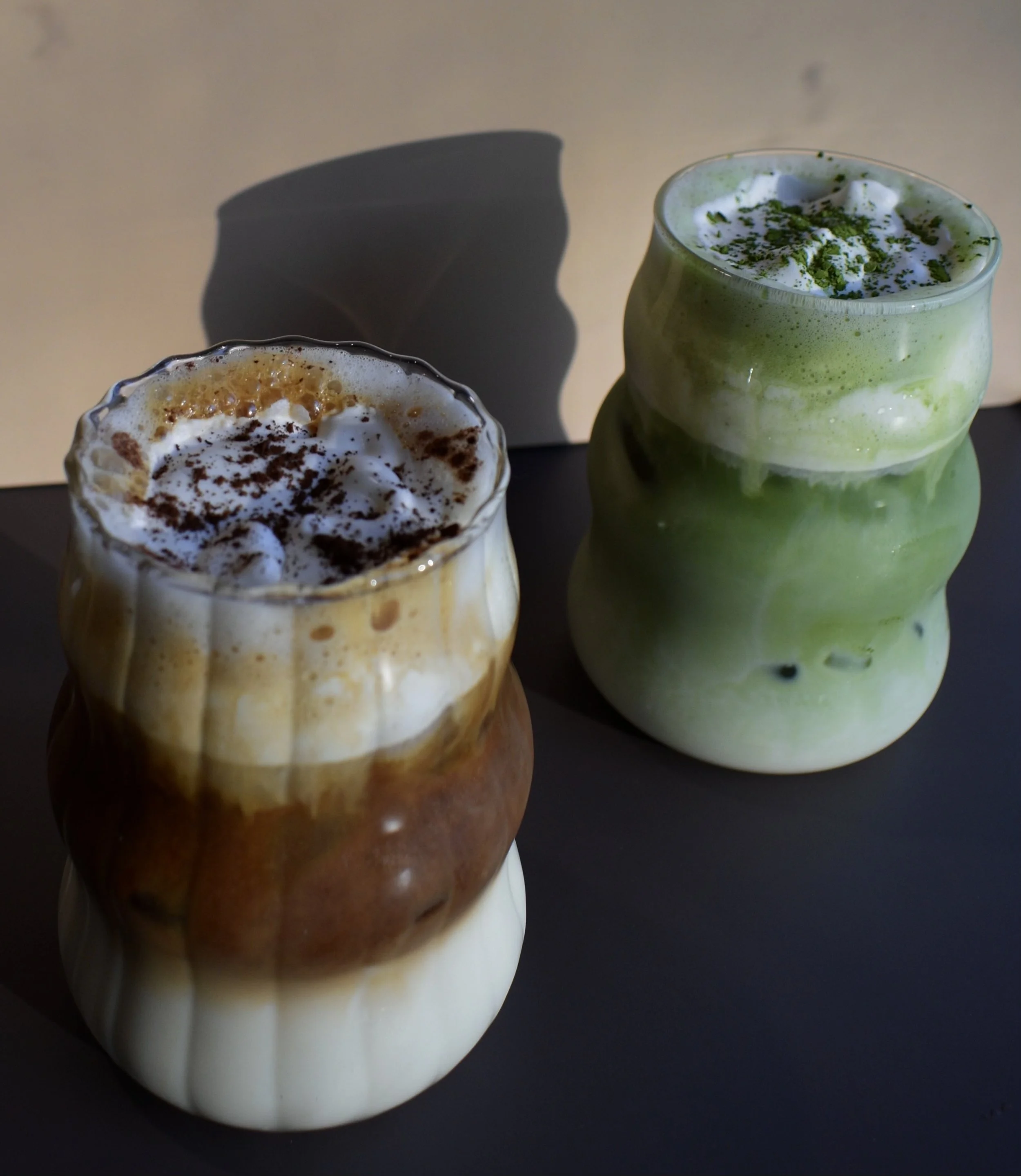 Two layered beverages in unique, wavy glass cups. The drink on the left appears to be a coffee or caramel-flavored drink topped with whipped cream and cocoa powder. The drink on the right is a green tea or matcha latte topped with whipped cream and green powder.