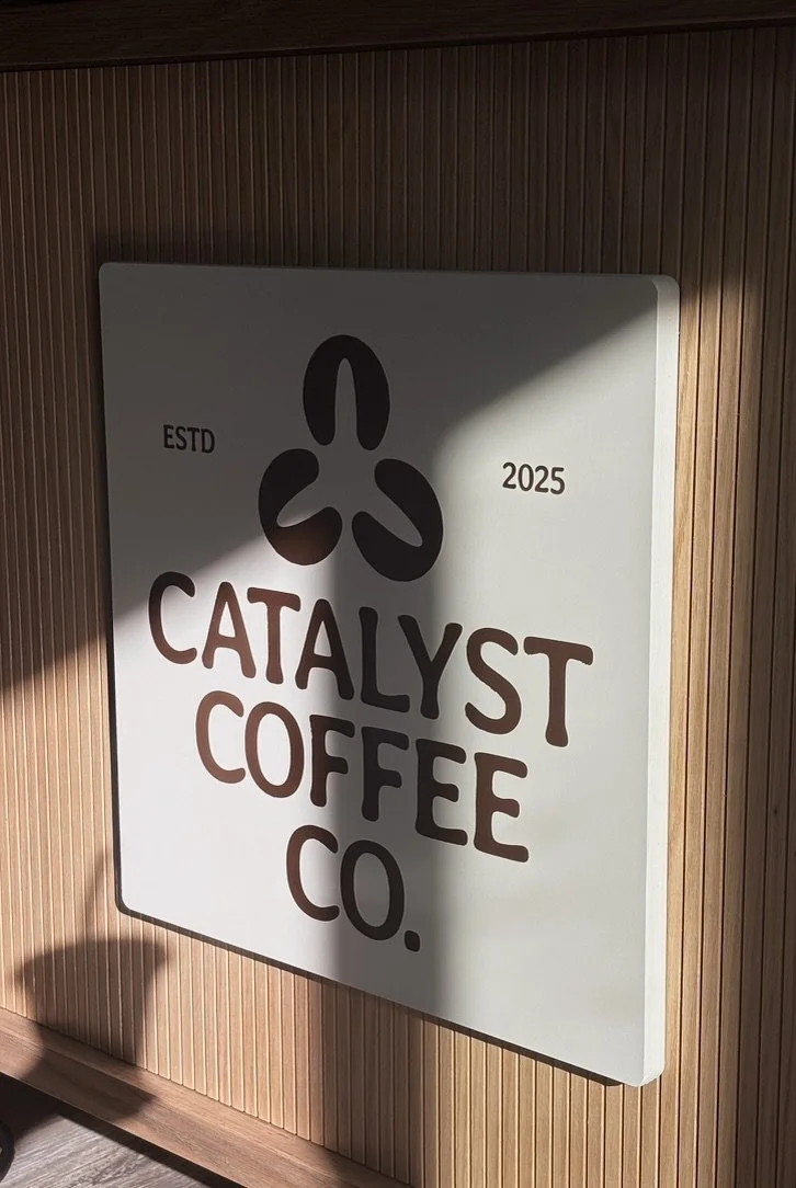 Sign for Catalyst Coffee Co. with a black logo resembling a stylized flower or plant, mounted on a wooden wall.