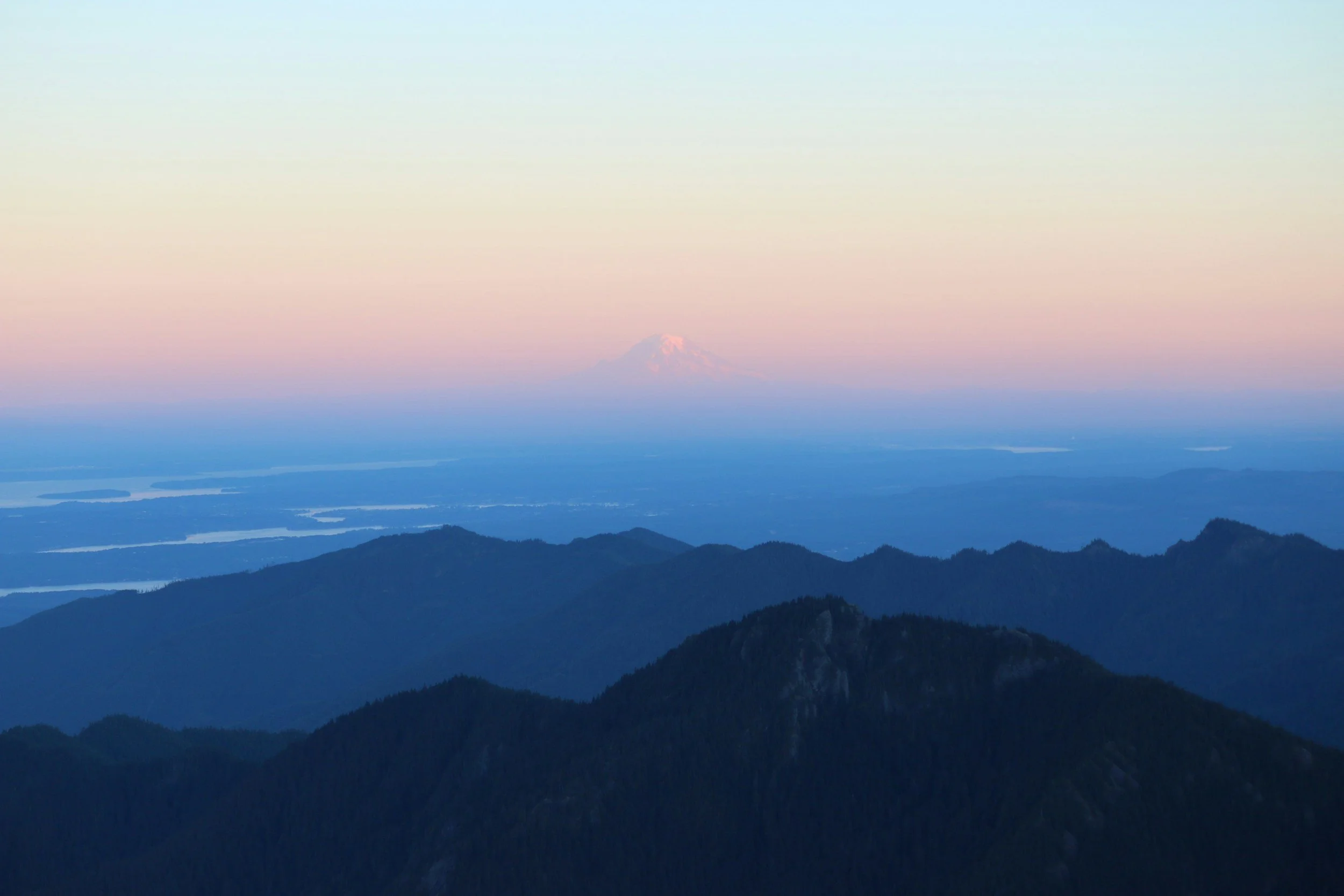 A scenic view from a mountain, showing multiple mountain ridges in the foreground and a snow-capped mountain in the distance, with a colorful sky during sunset or sunrise.