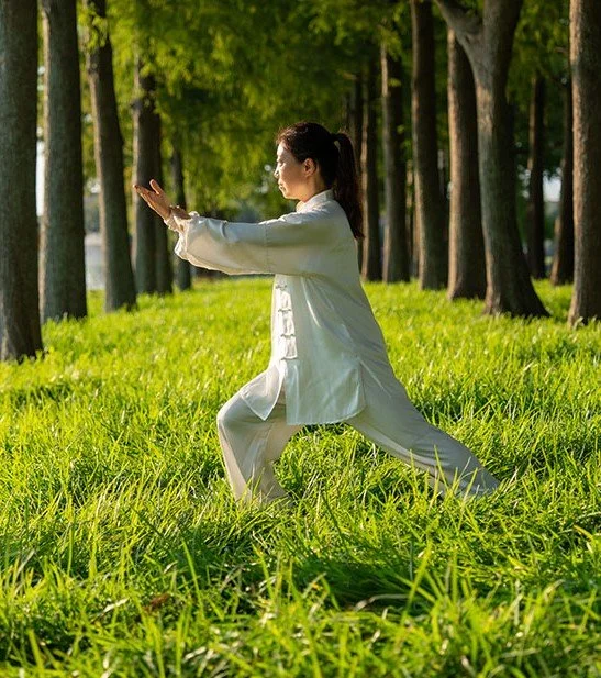 Shifu Fong in white traditional martial arts outfit practicing Tai Chi outdoors in a grassy field with trees.