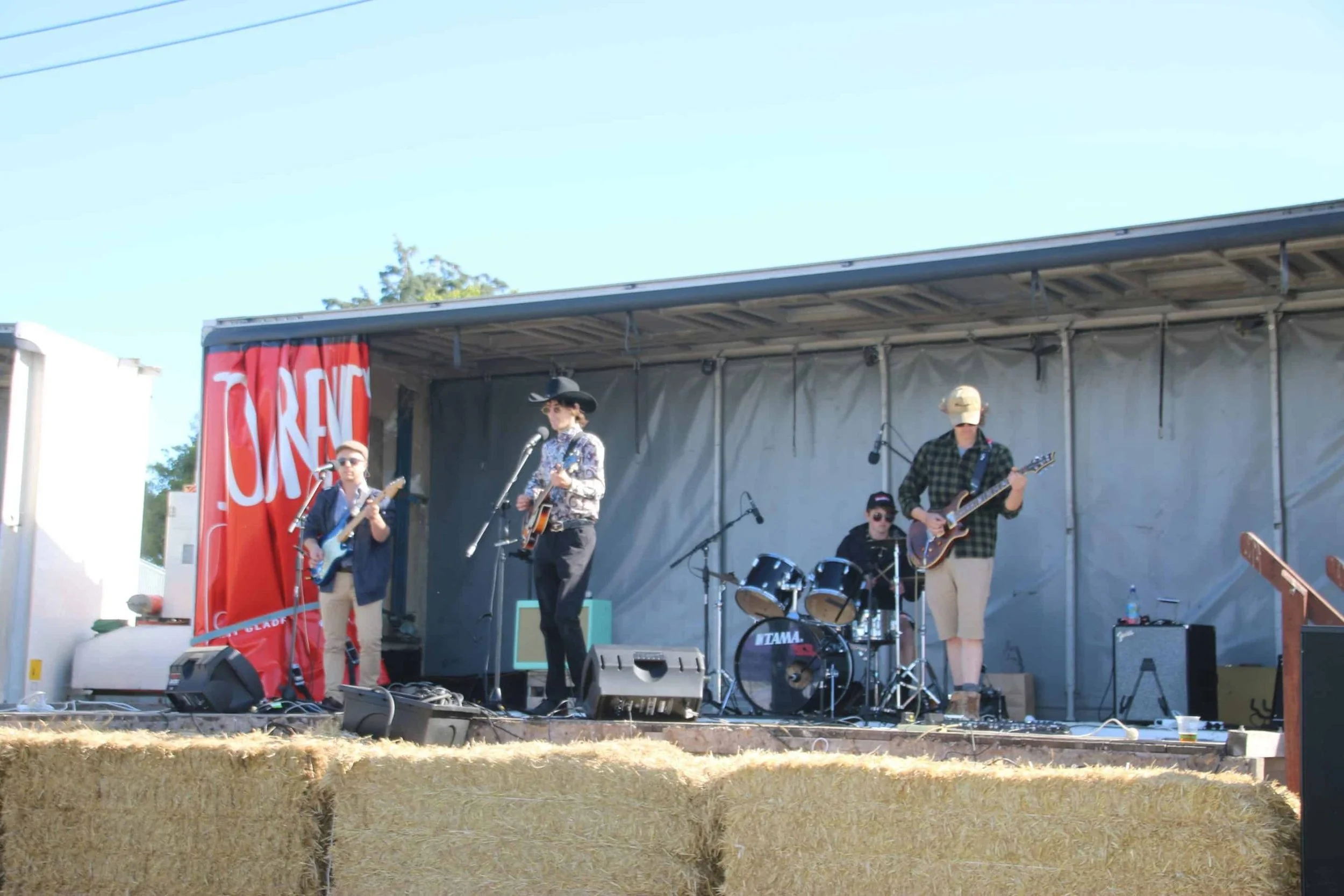 A band of five musicians performing on an outdoor stage with a red banner that says "CUREN". They are playing guitars and drums, with microphones set up for singing. The stage is surrounded by hay bales and there is a clear blue sky overhead.