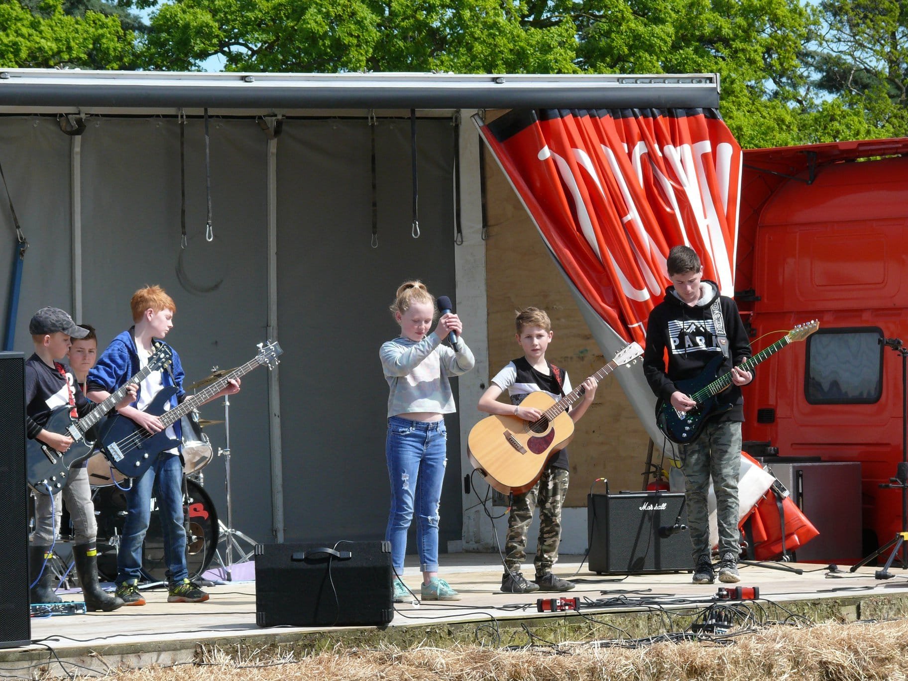 A group of children performing on an outdoor stage, playing guitars and singing, with a girl holding a microphone. The stage has a red truck trailer with a curtain and a canopy, and is surrounded by trees.