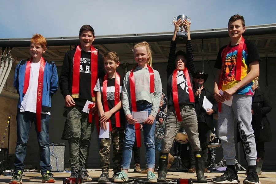 Children standing on stage with awards, medals, and papers, celebrating a victory, with a woman dressed in black and holding a microphone in the background.