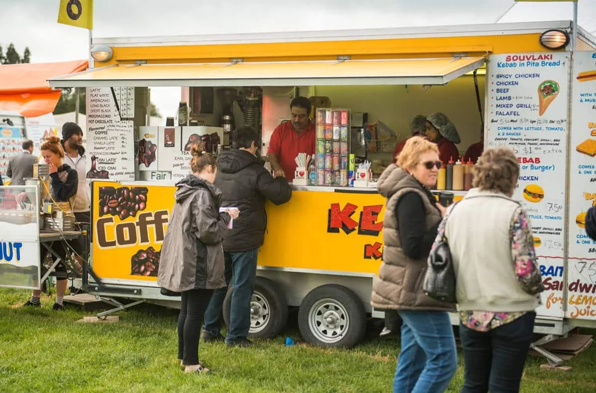 People standing in line at a yellow food stand selling coffee and drinks, with a menu board and other snack stands in the background.