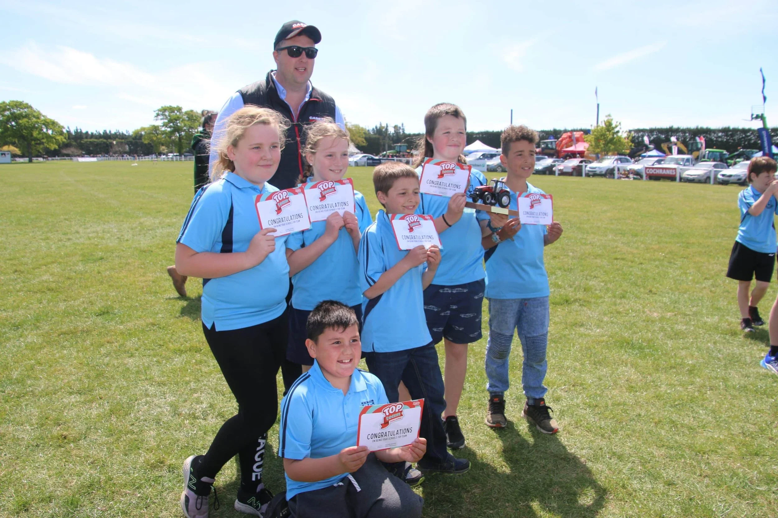 A group of children in blue sports shirts holding certificates, standing on a grassy field at an outdoor event, with a man standing behind them. Some kids are kneeling or standing, and others are smiling, holding trophies or awards.