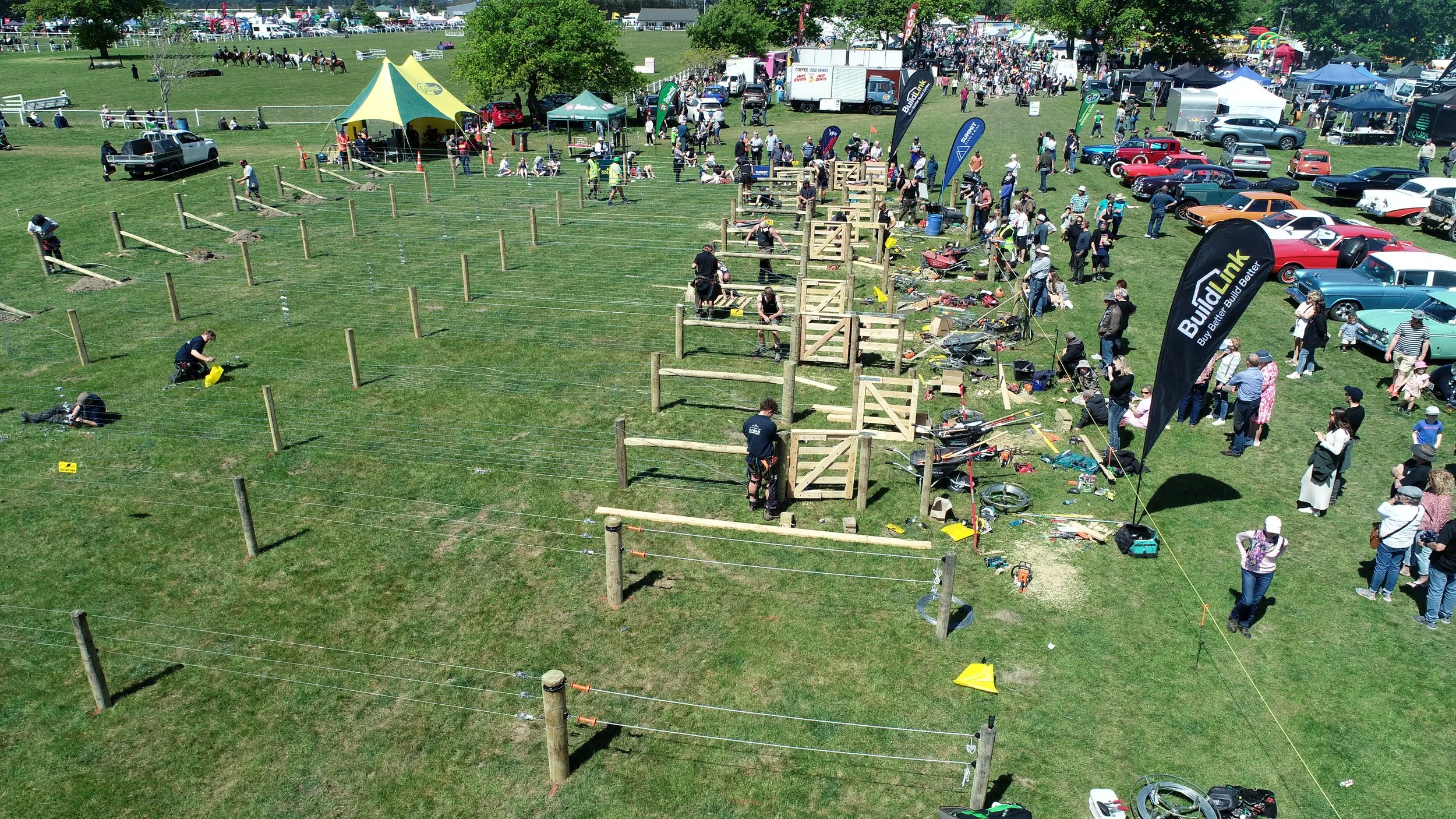 An outdoor event or festival with a crowd of people gathered around a construction project, involving building a wooden fence or structure. There are classic cars parked nearby, tents, flags, and a horse riding activity in the background.