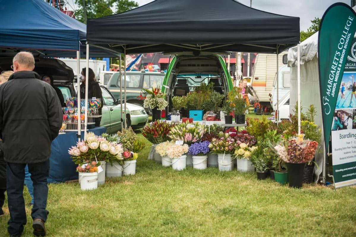 Flower vendor booth at an outdoor market with various flowers in buckets and potted plants, surrounded by tents and visitors.
