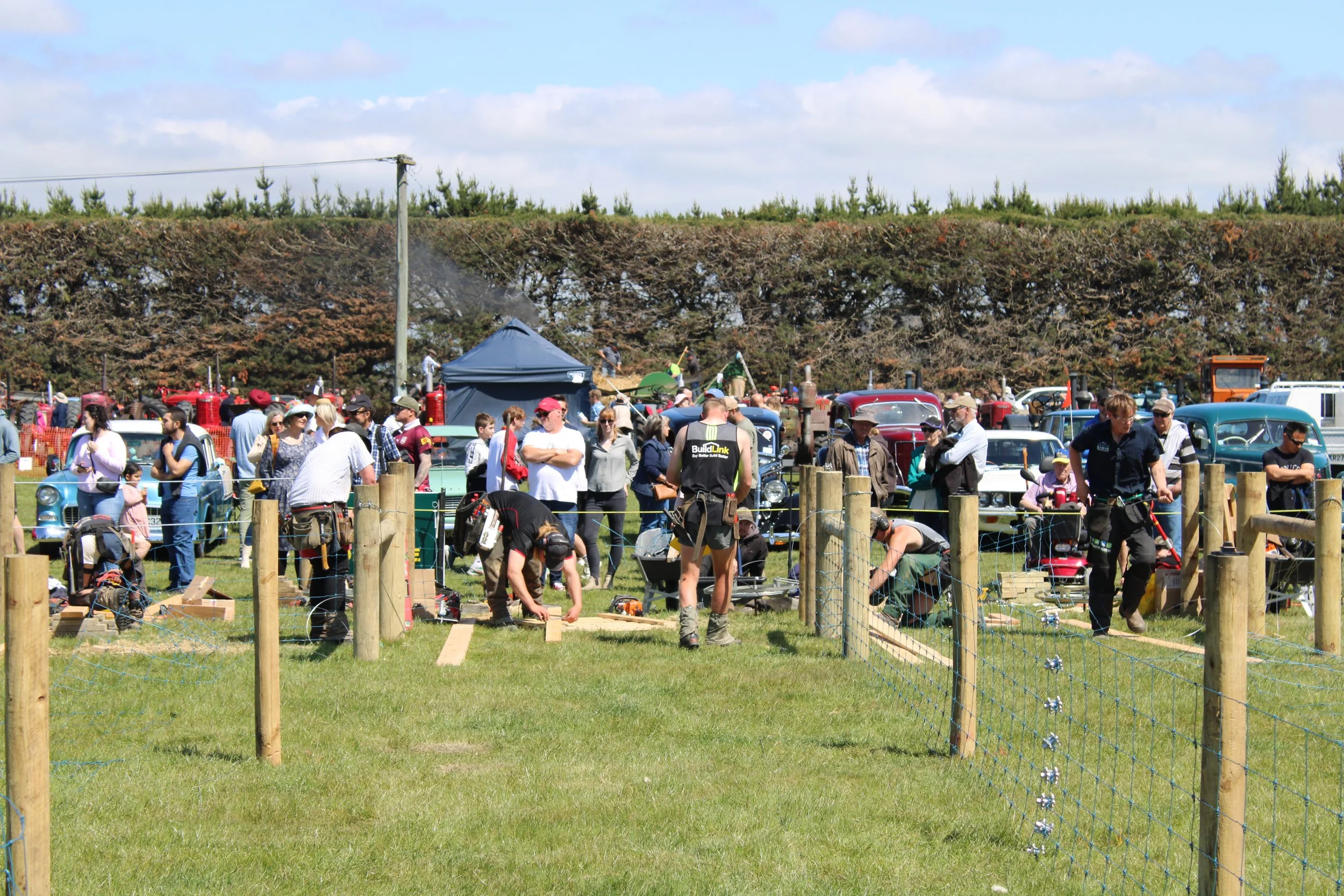 A busy outdoor event with people gathered around vintage trucks and tractors. There is a tent and a group of people, some observing and some working. The event is the Ellesmere A&P Show scheduled for Saturday, October 18th, 2025, at Ellersmere. The event features a fencing competition called 'Down to the Wire'.