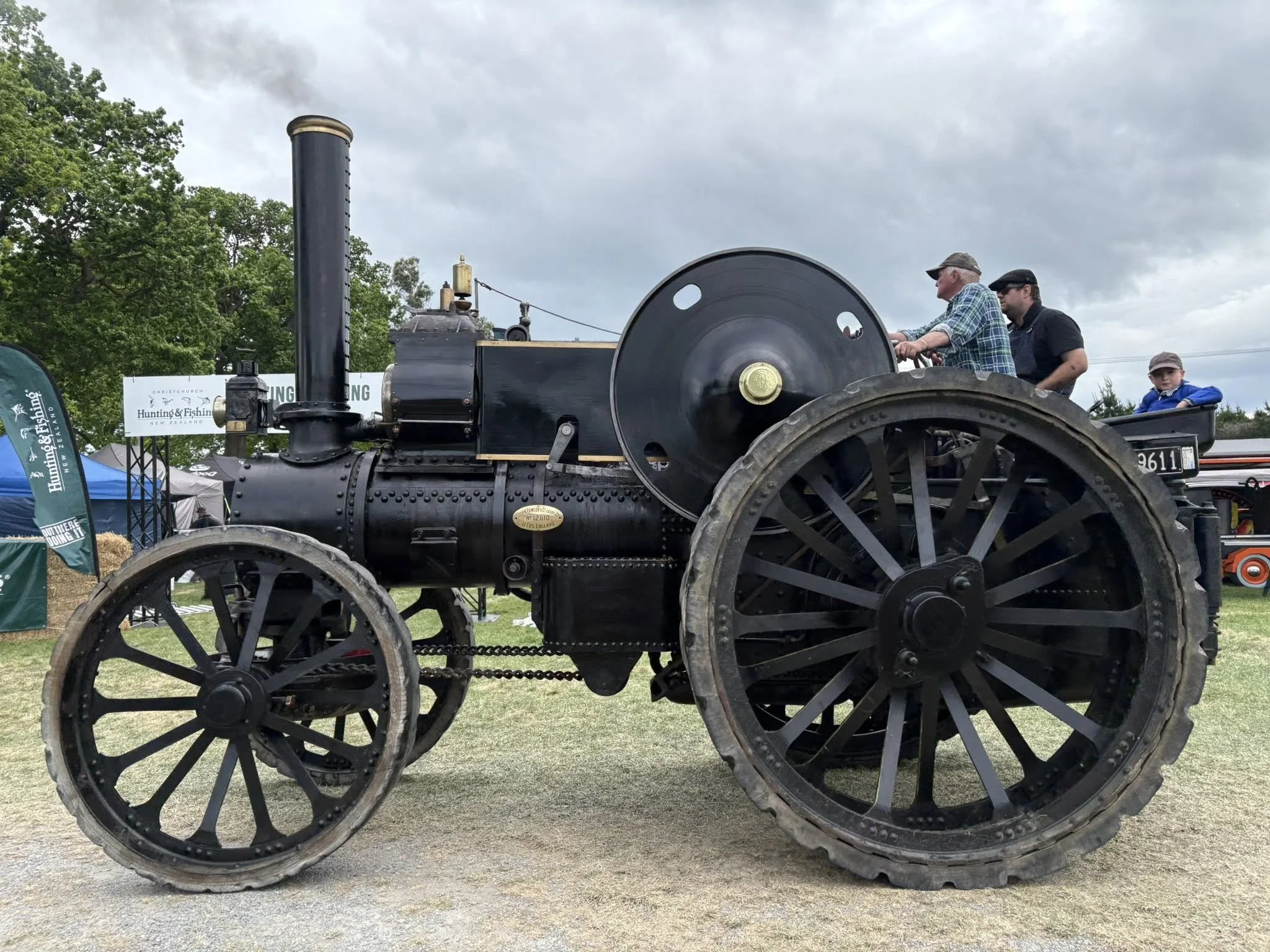 A large, black, vintage steam-powered tractor with big wheels, displayed outdoors at a fair or exhibition. Two men and a boy are sitting on or near it, under an overcast sky. There are tents and banners in the background.