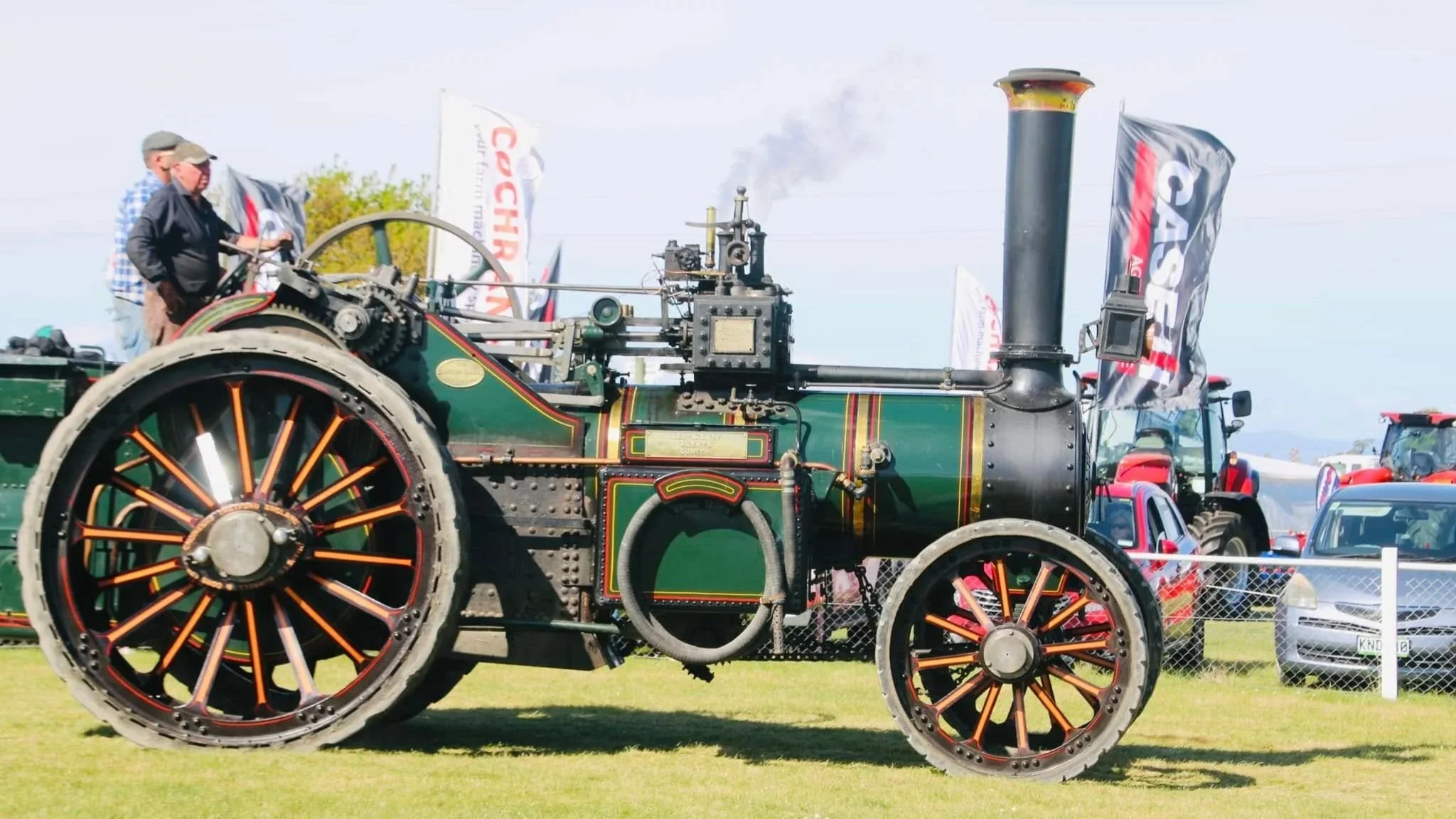 A vintage steam-powered traction engine with large, spoked wheels on a grassy field, with flags and people nearby at an outdoor event.