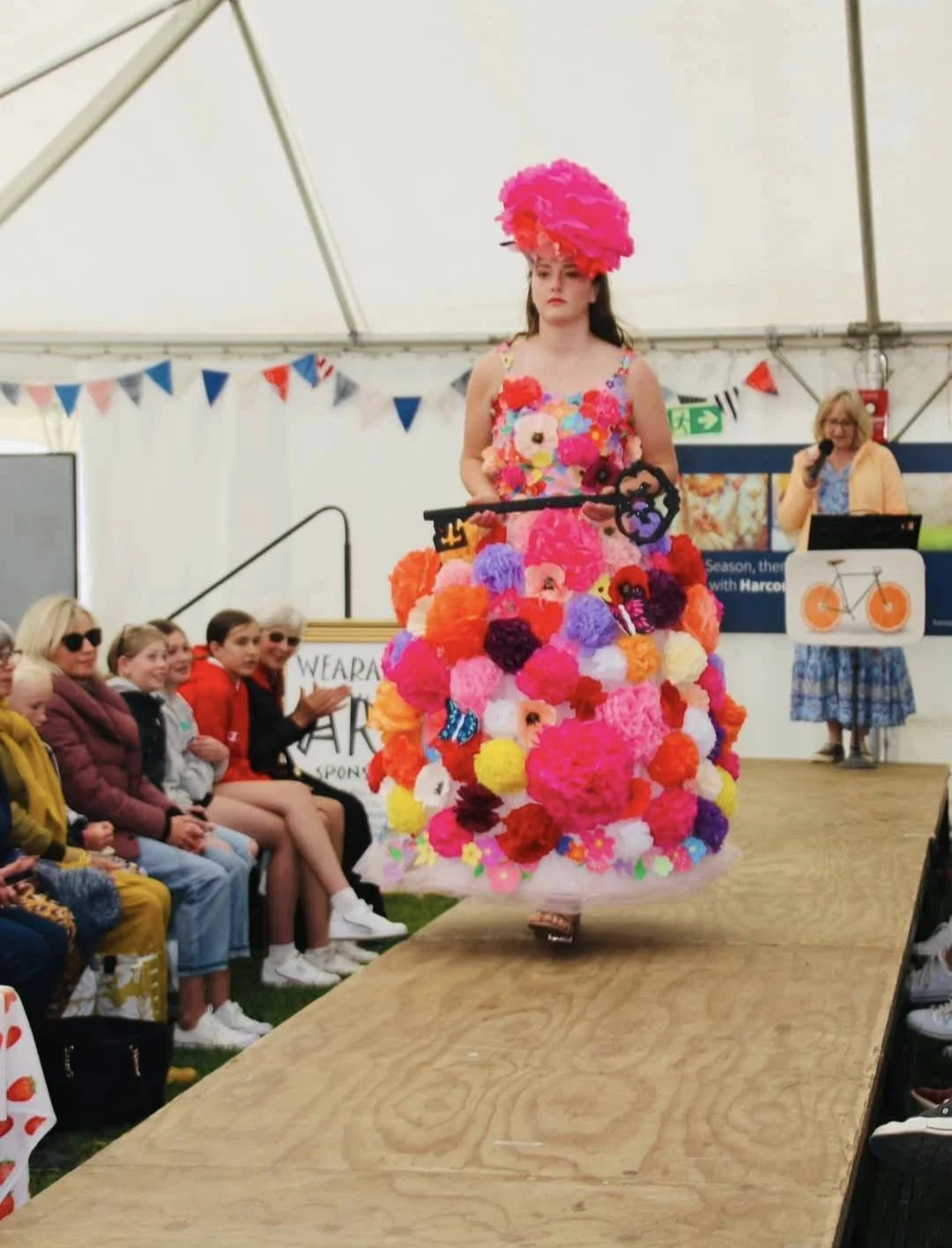 A young girl models a dress made of colorful pom-poms at a fashion show inside a tent, with spectators seated on the side.