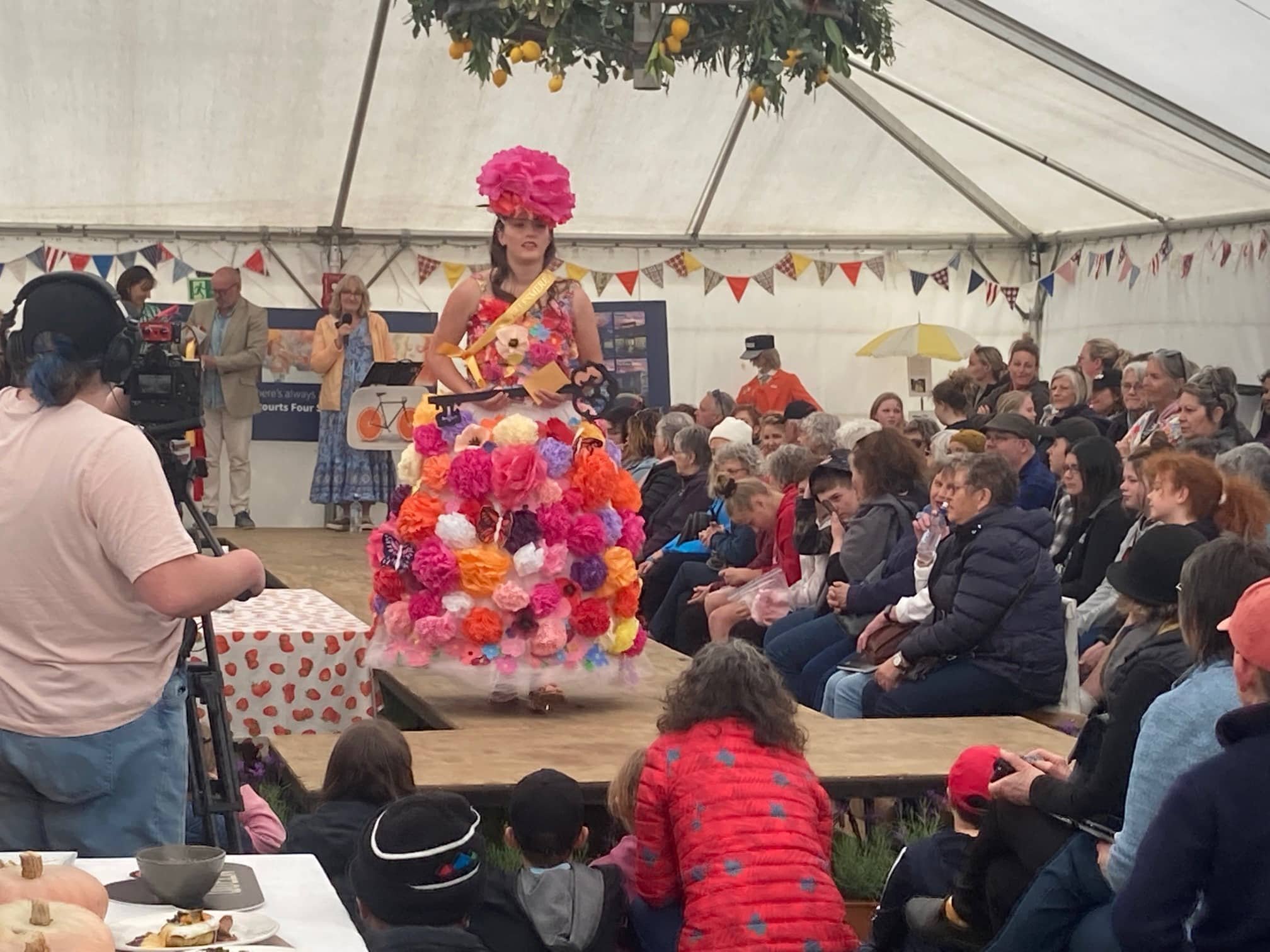 A woman dressed in a floral dress with a sash and wearing a large pink paper flower hat stands on stage in a large white tent, performing or speaking, with an audience seated watching her. The stage is decorated with bunting and orange trees.