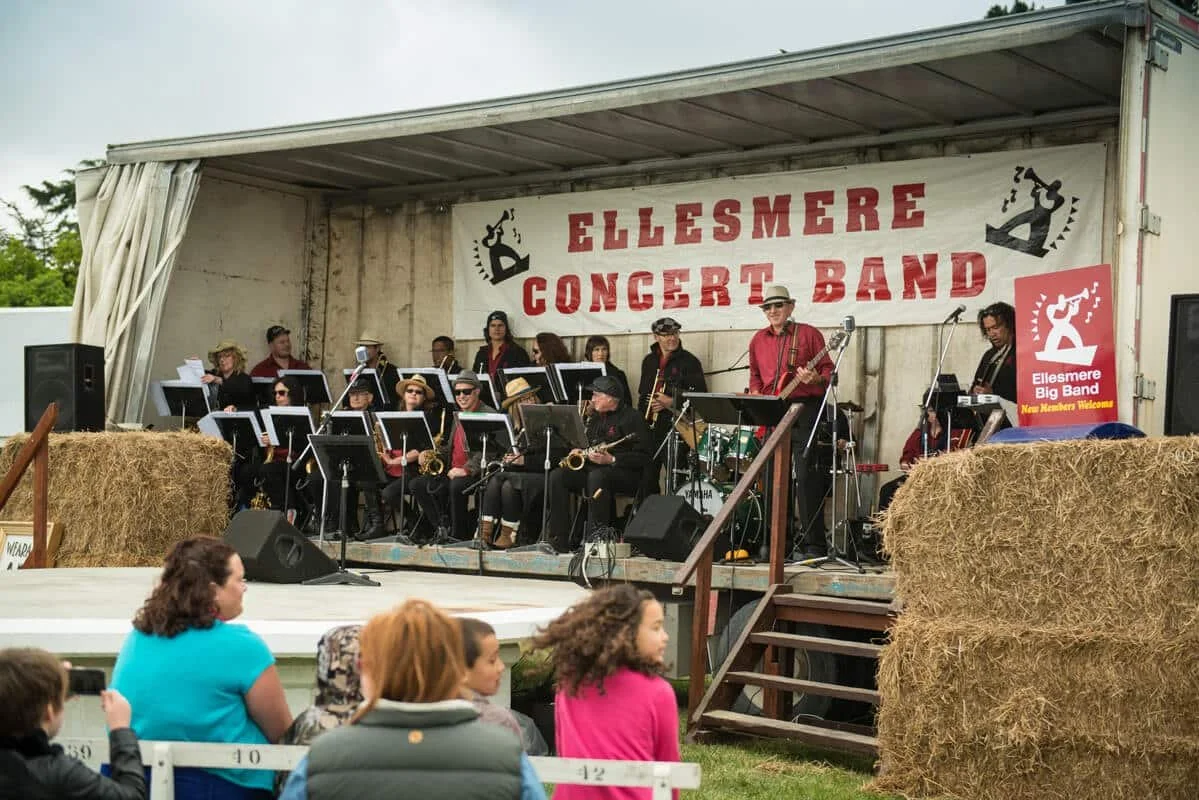 A band performs on an outdoor stage decorated with hay bales, labeled 'Ellesmere Concert Band.' Several musicians are seated with instruments, and some are standing. Audience members sit and watch in front of the stage.