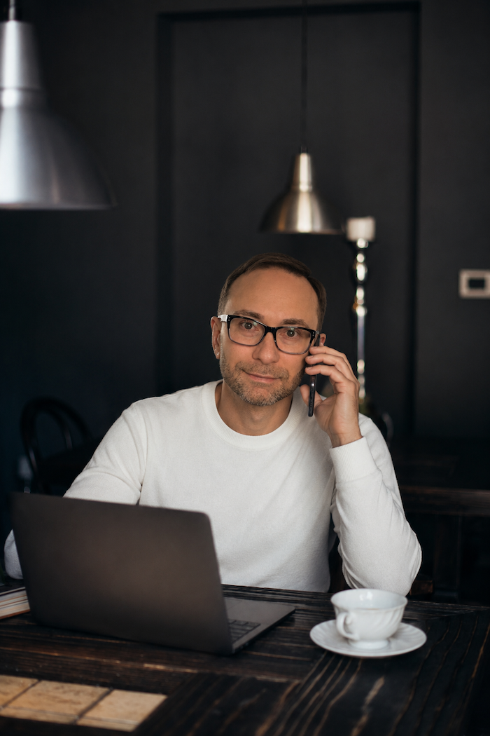 Alex Krasovsky Health Coach, Nutritionist sitting at a dark wooden table with a laptop, talking on a phone, and a cup on a saucer in front of him.