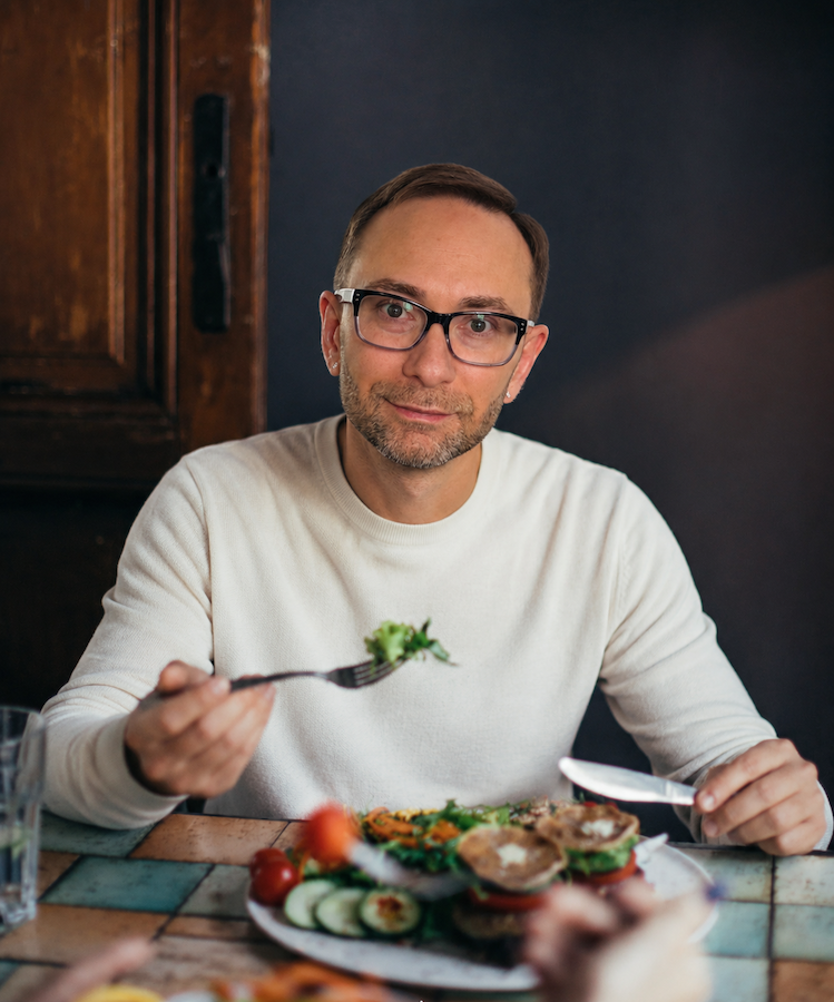 Alex Krasovsky Health Coach with glasses and a beard, wearing a white sweater, sitting at a table with a plate of food in front of him. He is holding a fork with a piece of vegetables and looking at the camera.