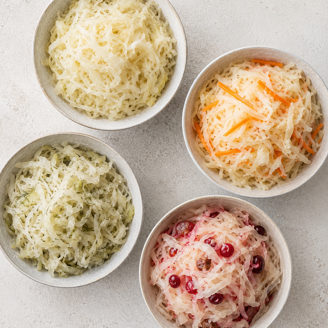 Four bowls of homemade sauerkraut variations with cabbage, carrots, dill, and cranberries on a light stone background.