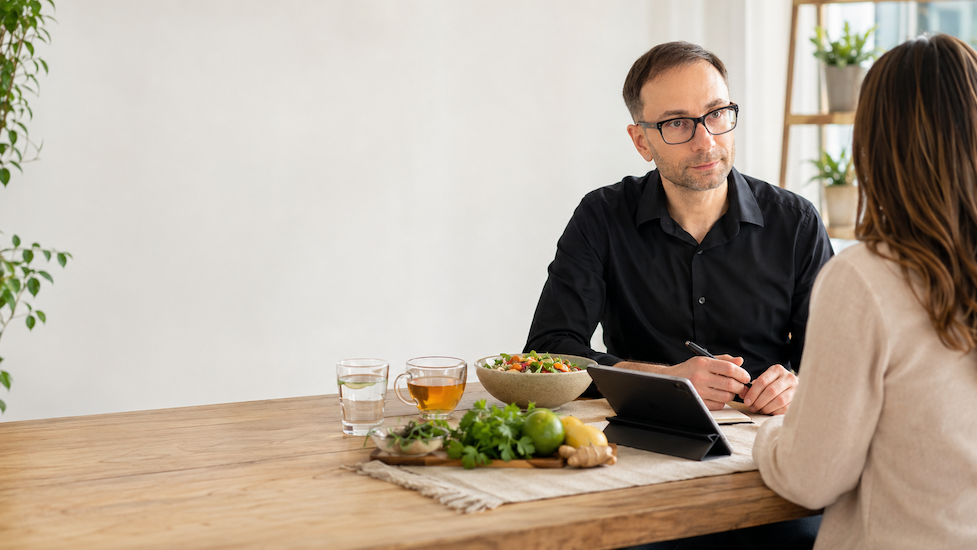 Alex Krasovsky Health Coach and woman sitting at a wooden table having a discussion in a bright room. On the table are a tablet, a bowl of salad, a glass of water, a cup of tea, and fresh vegetables and herbs.