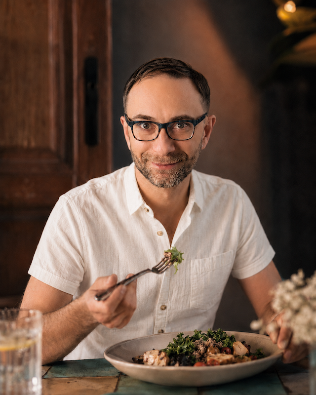 Alex Krasovsky Health Coach  with glasses and a beard wearing a white short-sleeved shirt sitting at a table holding a fork with food, smiling at the camera, with a plate of food in front of him and a glass of water nearby.