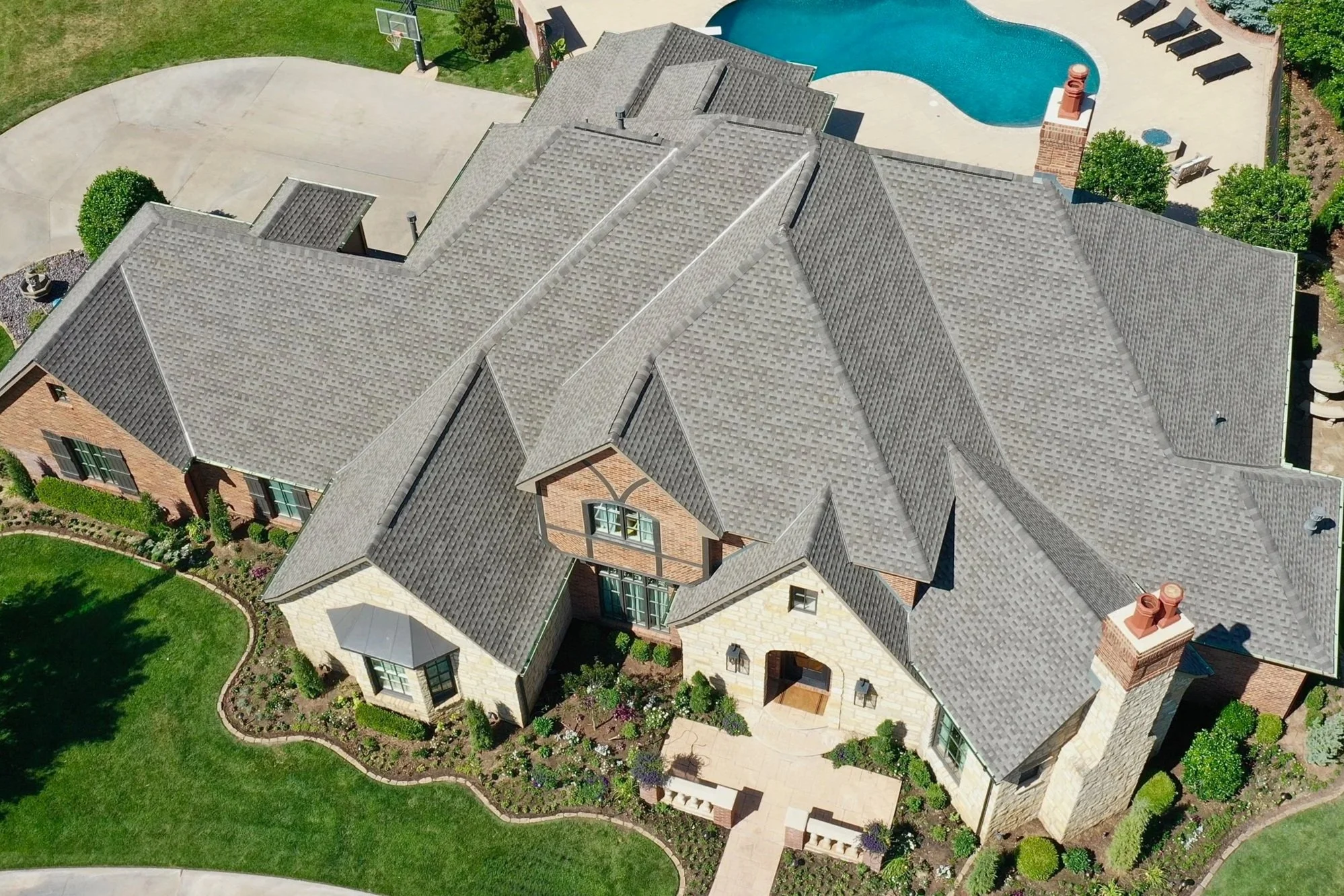 An aerial view of a large house with multiple gray shingled roofs. The backyard features a kidney-shaped swimming pool with lounge chairs and a brick chimney. The yard is landscaped with green lawns, trees, and flower beds.