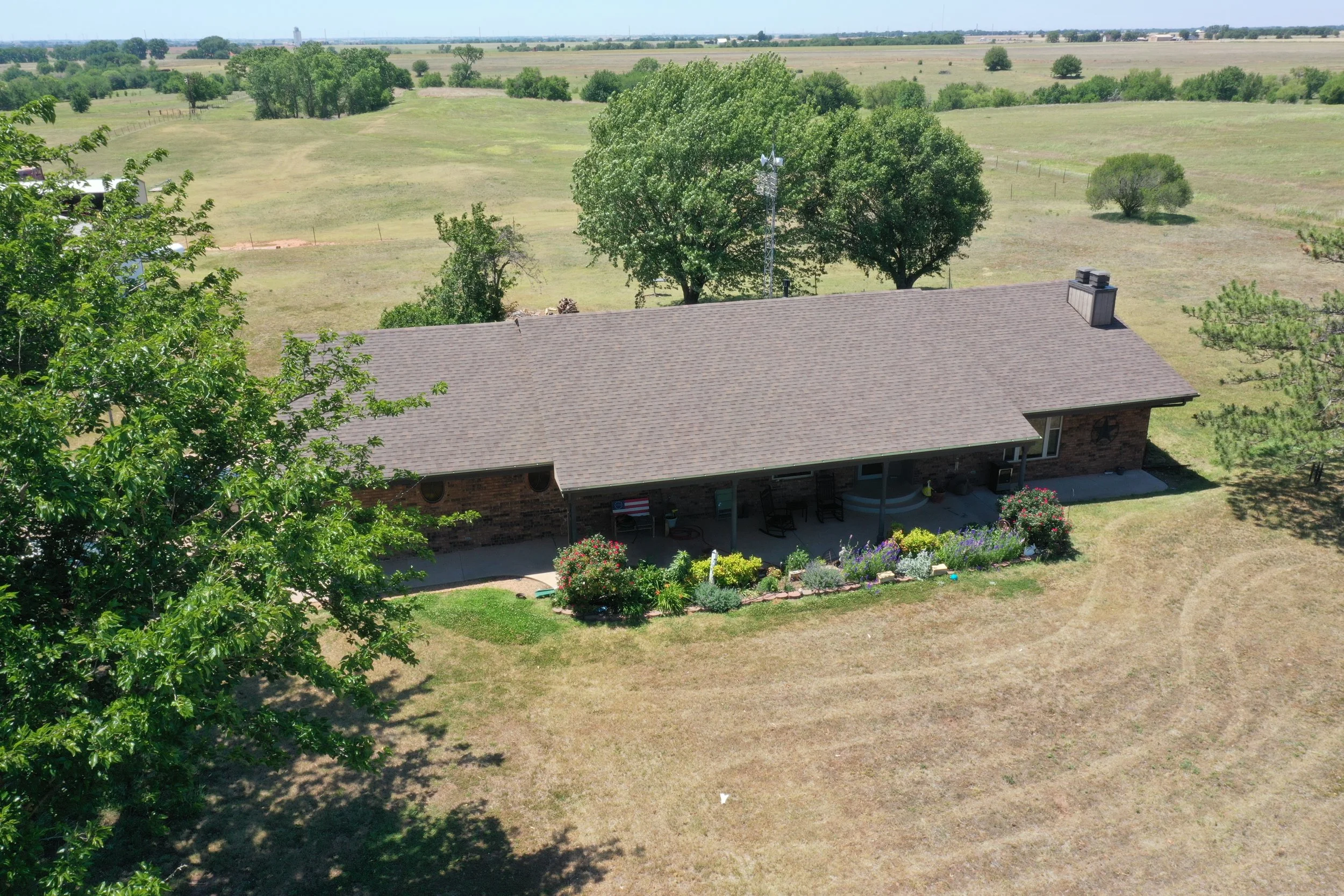 Aerial view of a rural homestead with a brown roof, surrounded by trees and a green lawn, with open pasture and a clear blue sky in the background.