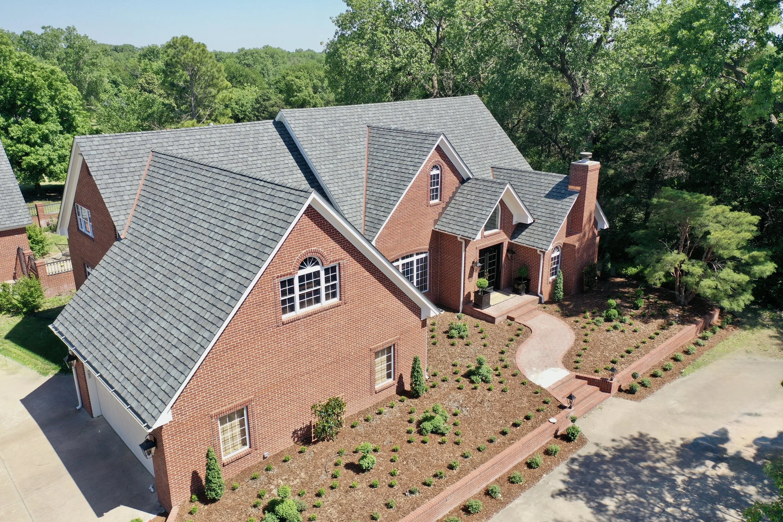 Aerial view of a red brick house with gray shingled roofs, surrounded by green trees and landscaped garden with small bushes and a pathway.