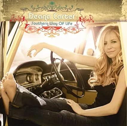 A woman with long blonde hair sitting inside a vintage vehicle, with her left arm resting on the steering wheel. The image has a warm, sepia-toned filter. There is a decorative banner at the top with the text "Deana Carter" and "Southern Way of Life."