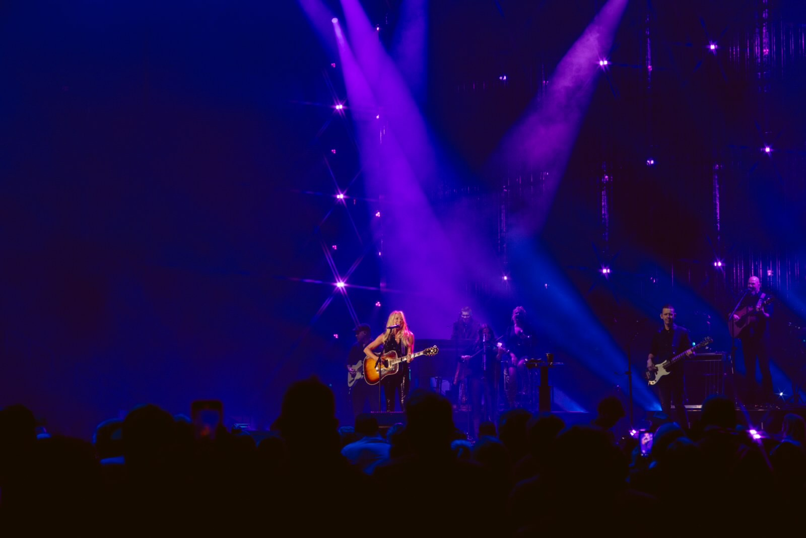 Musicians performing on a stage with purple and blue lighting, featuring a lead singer with a guitar and backup band members.