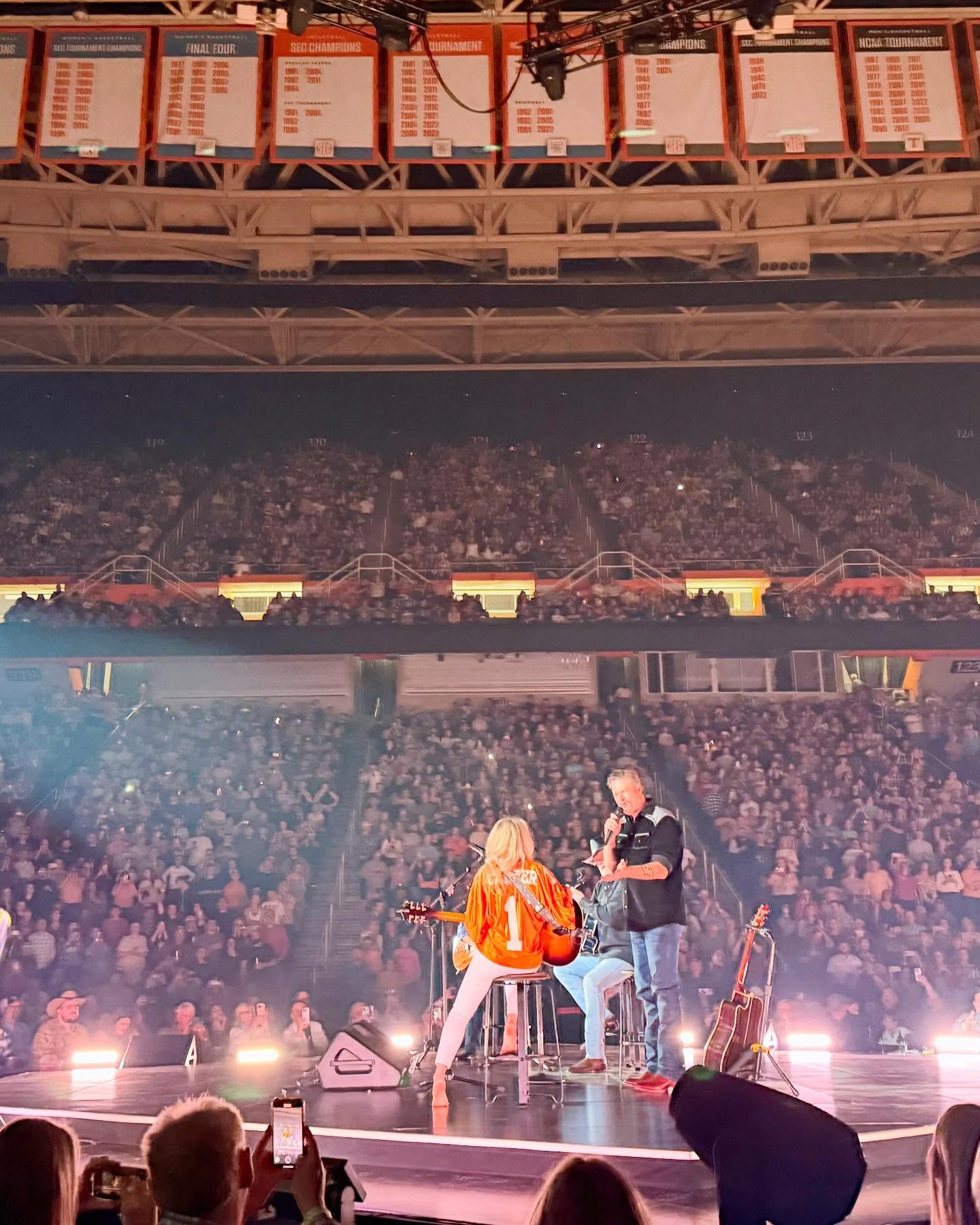 A person wearing a sports jacket giving a speech on stage while another person in a sports jersey sits beside them, holding a guitar. The stage is filled with audience members, and there are multiple tiers of seating above with banners listing sports championship years.