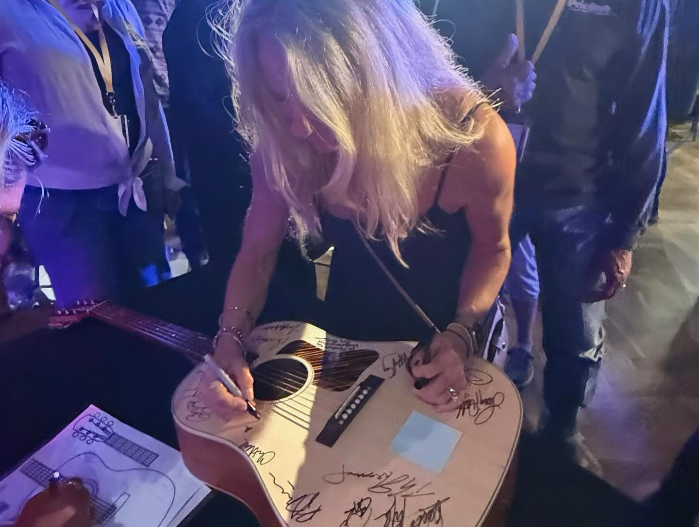 Woman with long blonde hair signing a decorated acoustic guitar at a crowded event, surrounded by people.