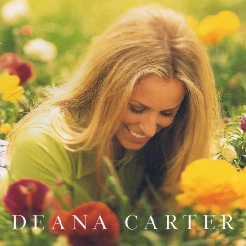 A woman with long blonde hair smiling and looking down among colorful flowers in a garden.