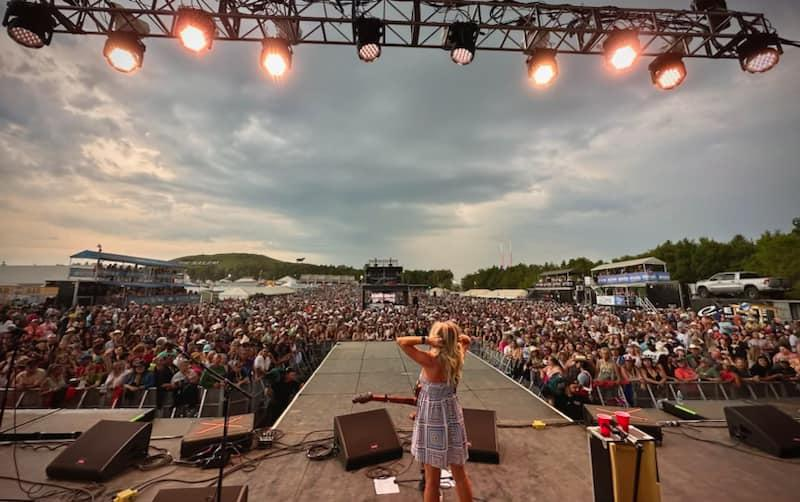 A woman on stage at an outdoor concert venue facing a large crowd with tents and trees in the background during a cloudy sky.