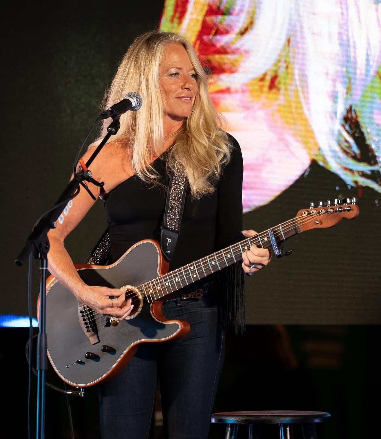 A woman with long blonde hair playing an acoustic guitar on stage, with a microphone in front of her and a colorful digital screen in the background.