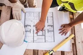 Hands pointing at a blueprint of a building, with a white safety helmet and construction tools on a table.