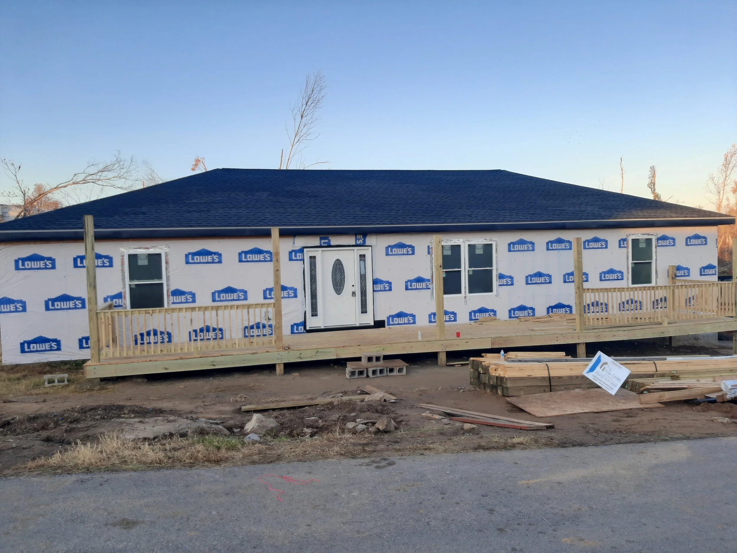 A house under construction with siding partially installed, a deck with railing, and construction materials on the ground.