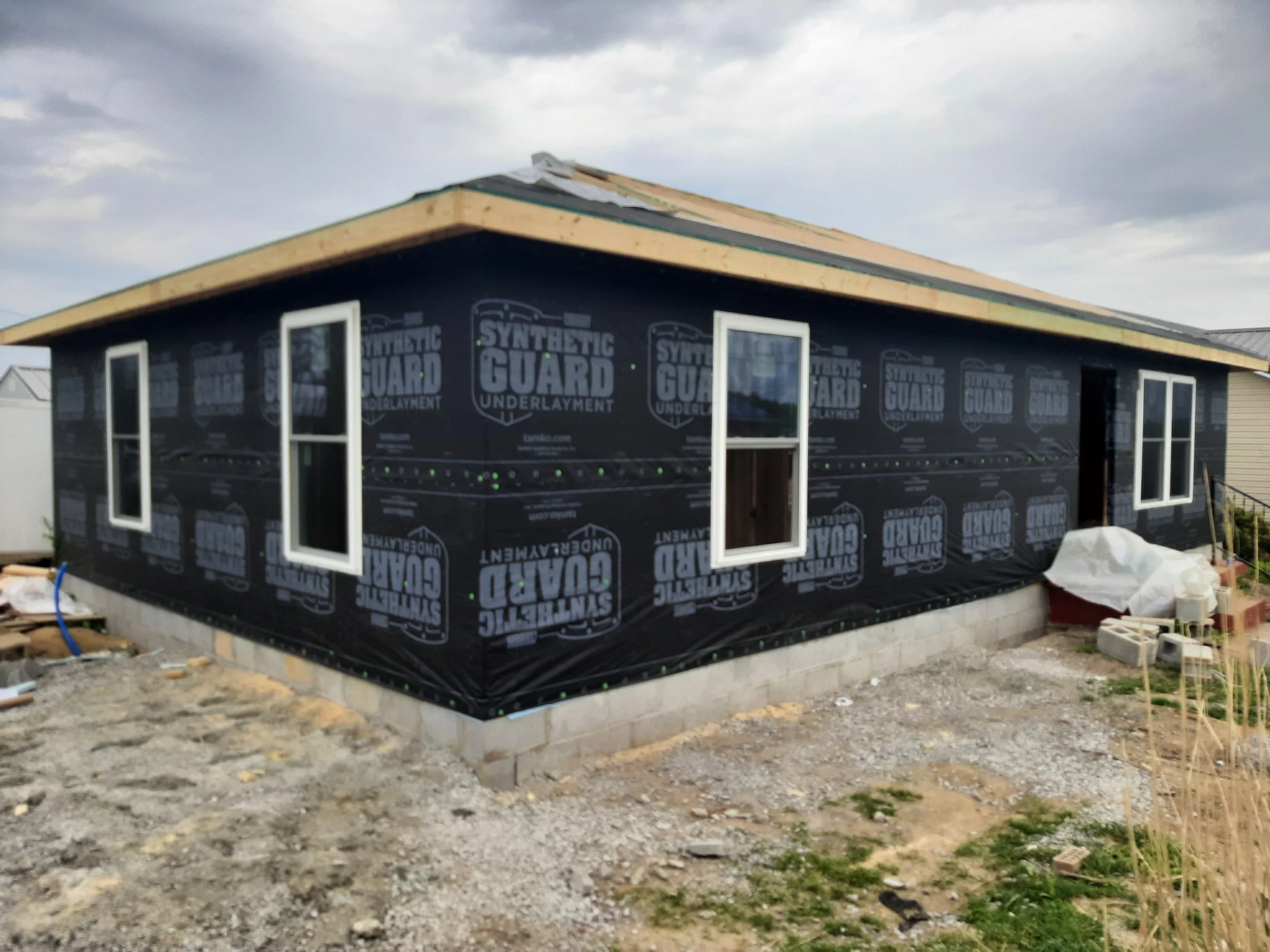 A house under construction with black underlayment and four windows installed. The roof is partially unfinished, and construction materials are scattered around on a gravel and dirt ground.